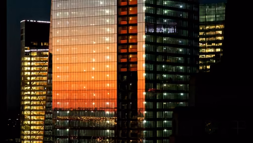 Glass office towers at dusk, the sunset reflected in one facade and the floors lit