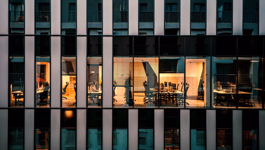 Office facade at blue hour, lit meeting rooms and desks behind the glass