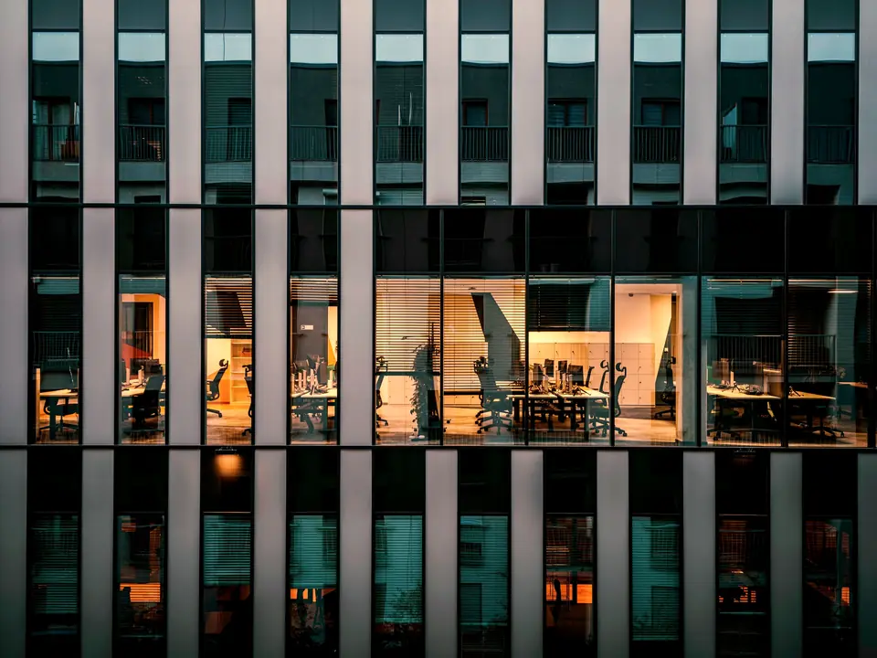 Office facade at blue hour, lit meeting rooms and desks behind the glass