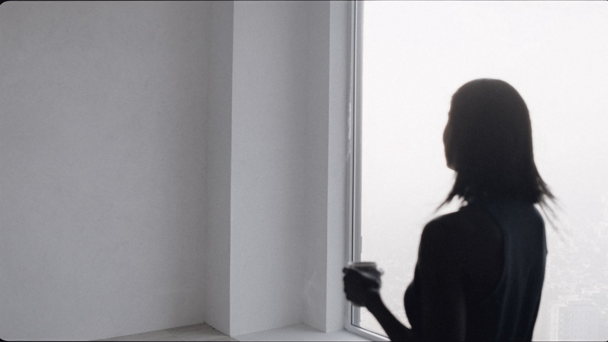 A woman holding a coffee cup looks out of a high-rise window over the city