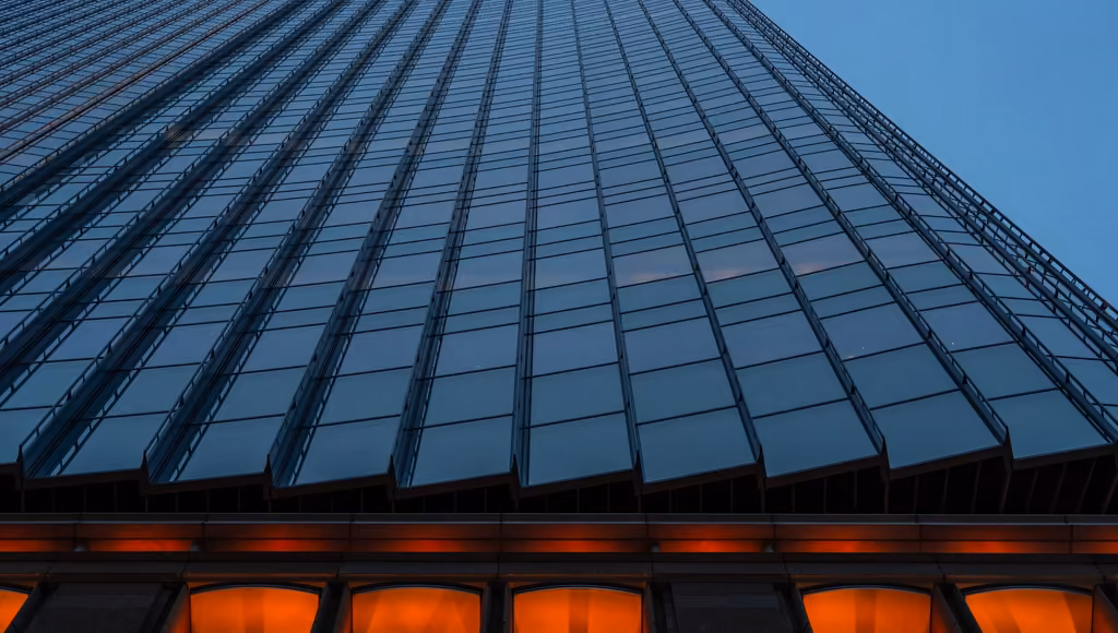 Glass curtain wall of an office tower at blue hour, its lit base glowing below
